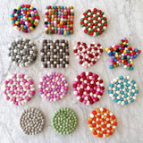 A handmade gray round felt ball placemat, crafted from individually hand-stitched organic wool felt balls, rests among other wool felt placemats on a marble surface. Made with 100% organic lamb wool ethically sourced from New Zealand, this unique felt trivet adds a touch of craftsmanship to any setting, ideal for protecting surfaces.