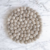 A round gray felt ball placemat, doubling as a pot trivet, rests on a clean white marble surface. It is densely packed with hand-stitched organic wool felt balls, creating a textured surface with subtle variations in gray tones. Ethically handmade by Nepalese female artisans, this handcrafted felt placemat adds a cozy, rustic elegance to home decor.