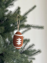 A brown needle felted American football ornament, part of an American Popular Sports Ornament Set, hangs from a Christmas tree branch. This 3-inch 3D wool felt ornament features detailed white stitching, adding a sporty touch to holiday decor.