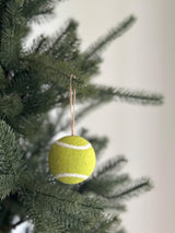 A needle felted tennis ball ornament hangs on a green Christmas tree branch. This round, bright yellow ornament features distinct white curved lines, mirroring a real tennis ball. Crafted from 100% needle-felted wool, it measures 3 inches in diameter and is suspended by a jute string, creating a festive, sporty display.