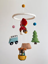 A wool felt camping baby mobile is suspended against a plain white background, creating a calm and whimsical atmosphere. It features a white felt ring from which colorful natural New Zealand sheep wool felt elements hang, including a blue van, a green pine tree, a brown tent, a yellow backpack, a campfire, and a bear-adorned kayak. Each element has a soft, tactile impression and a playful, handmade quality.