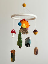 A handcrafted wool felt camping baby mobile is displayed, featuring a white felt ring suspending various camping-themed figures. Crafted from natural New Zealand sheep wool, these include a cozy campfire with orange and yellow flames, a green pine tree, a charming blue minivan with black wheels, and a green, brown, and yellow hiking backpack. A yellow felt ball tops the mobile, completing the outdoor adventure theme.