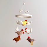 A colorful wool felt baby mobile hangs from the ceiling, featuring five vibrant chickens in playful poses against a plain white background. The chickens are handcrafted from organic New Zealand sheep wool and eco-friendly dyes, showcasing unique designs and shapes.