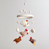 A baby mobile, made from wool felt, hangs from the ceiling against a neutral background. The mobile features five colorful chickens with different patterns: brown, white, and yellow. Each chicken is attached by a string and appears to float in mid-air, surrounded by small clouds made from soft material.