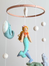 A baby mobile featuring a central mermaid figure and various sea creatures suspended above, made of wood with spherical beads and a soft design. The mobile is designed for a baby's room, evoking the tranquility of nature, particularly the ocean.