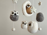 A baby mobile with an owl theme, featuring three felt owls hanging from a wooden frame, each with different expressions and colors. The mobile is surrounded by small felt balls resembling stars or clouds, attached to the frame by cotton strings. Made of wool felt, this charming mobile promises endless fun for your little one.