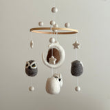 A charming wool felt owl baby mobile, suspended from the ceiling by soft cotton thread, features five owl figures with intricate details and fluffy texture. The wooden frame and colorful felt pieces create a whimsical theme, perfect for a child's room.