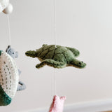 A wool felt sea life baby mobile is presented against a light, airy backdrop, featuring a central green wool felt sea turtle. The turtle has a detailed shell pattern and a soft, textured finish, appearing to float serenely on a thin cotton thread. It hangs from a sturdy wood frame, with other muted pastel wool felt sea creatures like a scallop and dolphin subtly visible, creating a cohesive underwater theme.