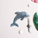 A close-up view of a Sea Life wool felt baby mobile, featuring a prominent soft blue wool felt dolphin, rests against a clean white background. The textured dolphin hangs by a thin white string with a small white felt ball. Subtly visible are other marine creatures, including a gray shark and a stingray, creating a calm underwater theme.