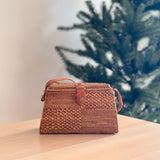 A woven rattan crossbody bag, Maya model, rests on a wooden table, its light brown color complementing the festive Christmas tree background with decorations. The bag features a centered leather snap clasp and an adjustable strap, showcasing its handcrafted quality with a textured surface made from natural materials like bamboo or rattan.