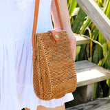 A person wearing a white top carries a woven rattan crossbody bag, made from natural fibers with textured surface and visible weaving patterns. The bag features an adjustable leather strap and snap closure, suitable for carrying small items like a cellphone. The background is a wooden staircase, suggesting casual outdoor use.