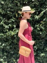 A woman stands outdoors, surrounded by lush green foliage, wearing a maroon sleeveless dress, wide-brimmed straw hat, and sunglasses. She holds a woven rattan crossbody bag with a leather strap, showcasing its oval shape and smooth material.