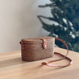 A woven crossbody bag crafted from Ata grass and real leather, resting on a surface in a cozy indoor setting. The rectangular shape features rounded corners, an adjustable brown leather strap for carrying over the shoulder, and a compact design. Made from environmentally friendly materials, this handcrafted piece exudes timeless elegance.