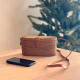 A woven crossbody bag, crafted from natural materials rattan or Ata grass, rests on a wooden surface. The textured finish features a small top handle and an adjustable strap that wraps around the body of the bag. The bag's natural texture and practical design evoke a cozy atmosphere, perfect for carrying personal items like a phone.