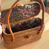 A petite rattan handbag, Catania, resting on a woven mat, showcasing its smooth finish and real leather handles. The bag features a unique Batik cotton lining and is made from eco-friendly materials.