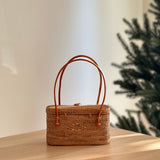 A woven rattan handbag with a smooth finish, resting on a wooden table against a festive Christmas tree backdrop. The bag features a snap closure and leather handles, showcasing its stylish and eco-friendly design.