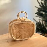 A woven handbag with a natural rattan handle sits on a wooden surface, surrounded by a Christmas tree backdrop and a section of wall. The bag's eco-friendly materials and unique design elements, such as batik lining and drawstring pouch, are highlighted. The overall mood is warm and inviting, suggesting a festive atmosphere.