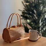 A wicker handbag with a guava-shaped design rests on a wooden surface next to a small cup, evoking a holiday atmosphere with a Christmas tree visible in the background. The bag features a distinctive bamboo-like pattern and a handle made from the same material.
