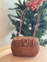 A person's hand holds a woven rattan crossbody bag with a real leather handle, resting on a wooden surface in an indoor setting. The light brown bag has an oval shape with a fitted lid featuring a leather hook closure and is made of natural materials like rattan or wicker.