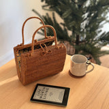 A woven rattan handbag, Bali Rattan Handbag - Kenia, rests on a table next to a smartphone displaying a book cover. The handbag features a textured surface, sturdy base, and short handles made from Ata grass, adorned with a Batik lining showcasing traditional Balinese patterns.