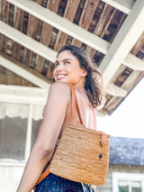 A woman stands outdoors, wearing a sleeveless top and blue jeans, holding a rectangular rattan backpack over her shoulder. The bag has a brown leather strap and appears to be made of natural materials with a simple design featuring a sleek arch shape and secure snap closure.