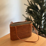 A Bali Rattan Shoulder Bag rests on a wooden surface, its oval shape at the top view showcasing sturdy leather handles for easy carrying. The bag's rattan material and refined touch evoke a sense of eco-friendliness and fair trade practices.
