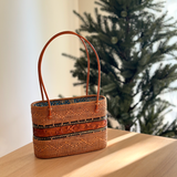 A handcrafted woven rattan tote bag rests on a wooden surface, showcasing its premium real leather handle adorned with intricate wood carvings. The snag-free finish ensures smooth handling, while the unique Batik cotton lining adds style and longevity.