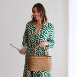 A woman stands in front of a white wall, holding a woven rattan handbag with a green and white pattern. The handbag is made from natural materials and features a textured design. It's being held by the woman, suggesting it might be used for carrying personal items or as a fashion accessory.