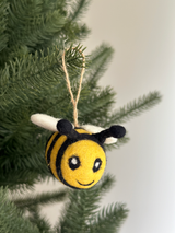 A cheerful felted wool bee ornament hangs from a lush green Christmas tree branch by a jute string, creating a festive atmosphere. This handmade felt bee ornament features a striped yellow and black body with small white wings and black antennae. Its face is adorned with black eyes and a smiling mouth, adding a whimsical touch to holiday decor.