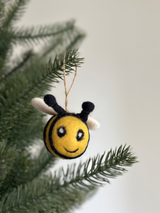 A charming felted wool bee ornament hangs gracefully from a green Christmas tree branch, set against a soft off-white backdrop. This unique holiday decoration features a bright yellow, round body with black stripes, a smiling mouth, small black antennae, and cream-colored wings. It is suspended by a jute string.