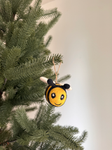 A cheerful felted wool bee ornament hangs gracefully on a lush green Christmas tree, set against a soft off-white wall. This round bee features bright yellow and black stripes, small black antennae, tiny white wings, and a heartwarming stitched smile with oval eyes. A jute string is visible at the top, facilitating its display.