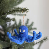 A wool felt blue octopus ornament hangs from a green pine branch against a soft white backdrop, exuding a whimsical and cheerful mood. This vibrant blue felt octopus ornament features eight curly tentacles, small black eyes, and a simple stitched smile. Made from fine wool felt, it offers a soft yet durable texture, ready for hanging by its jute string.