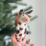 A handmade wool felt brown cow finger puppet is held up by a female hand against a blurred Christmas tree background. This charming puppet is primarily white with distinctive brown spots, ears, and horns. Its gentle expression is created with needle-felted details and a black stitched smile.