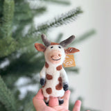 A unique, handmade brown cow finger puppet, crafted from wool felt, rests in the palm of a hand against a blurred Christmas tree background. This charming needle-felted puppet is predominantly white with brown spots and black hooves. It features intricate details like a gentle smiling expression, small ears, and textured horns, making it a delightful toy for farm-themed creative play.