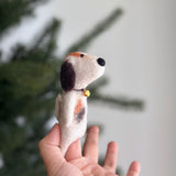 A playful handmade calico dog felt finger puppet is held by a female hand, set against a soft green Christmas tree and a white wall. This charming dog finger puppet is predominantly white with patches of brown, orange, and black, featuring a distinctive calico pattern and a small gold bell around its neck. Crafted from textured wool felt with needle-felted details, this unique finger puppet showcases its small, rounded shape.
