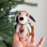 A handmade wool felt Calico Dog Finger Puppet is held in a female hand, set against a blurred Christmas tree backdrop. The unique felted finger puppet features a patchwork coat with white, brown, and orange markings. A small golden bell adorns its neck, adding to its charming appeal.