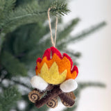 A cozy felt campfire marshmallow ornament hangs from a green Christmas tree branch. This unique Christmas ornament features layered red, orange, and yellow felt flames with three white felted marshmallows toasted over brown felt logs, detailed with spiral patterns, evoking a festive atmosphere.