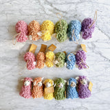 A set of six handcrafted felt sheep finger puppets is displayed in three rows on a marbled surface. Each unique wool felt sheep finger puppet, showcasing its craftsmanship, features a fluffy body, a small smiling face with white ears, and a loop with a tag. They come in soft pastel pink, orange, yellow, green, blue, and purple, creating a visually appealing spectrum.