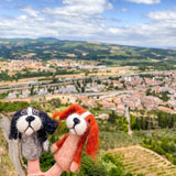 A handmade orange Cavalier King Charles Spaniel wool felt finger puppet is held up by a hand, set against a bright, airy backdrop of rolling hills and a sprawling town. This unique needle felted finger puppet showcases a white marking on its head and floppy ears, featuring a soft, textured appearance characteristic of its quality craftsmanship.