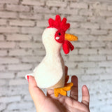 A handmade wool felt chicken finger puppet is held upright in a female hand with red nail polish, set against a blurred white brick background. This uniquely designed chicken features a white body, tiny black eyes, orange beak and feet, and a bright red comb and wattle, showcasing meticulous craftsmanship.