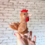 A brown wool felt chicken finger puppet is held upright by a female hand against a white brick background. This meticulously handmade chicken finger puppet features a brown body, a red felt crest and wattle, a light pink beak, and a small black eye. Its unique wing details are crafted with layered brown and light pink felt pieces, fanning out on its side.