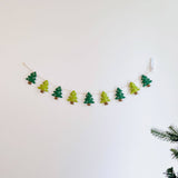A felt Christmas Tree Garland with jute thread hangs gracefully on a clean white wall, with a hint of a real Christmas tree in the lower right corner. This festive garland features alternating green and light green felt Christmas tree shapes, each adorned with embroidered white snowflakes and brown tree trunks. The jute thread provides a rustic, natural texture.