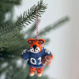 An Auburn Tiger wool felt ornament hangs playfully from a lush green Christmas tree branch, set against a soft, neutral background. This handcrafted wool felt Auburn Tiger ornament is vibrantly colored in navy and burnt orange, showcasing its characteristic stripes and team attire. Detailed needle felting brings out its bold and spirited character, suspended by a red and white cotton string.