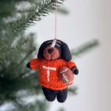 A handcrafted Tennessee Volunteers wool felt ornament hangs from a red and white cotton string against a soft, neutral background with hints of Christmas tree branches. This orange and white felt ornament showcases a bold 'T' design with visible hand-stitched details, perfectly capturing team spirit for any festive setting.