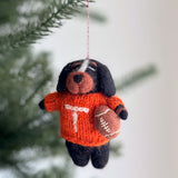 A handcrafted wool felt Tennessee Volunteers dog ornament hangs from a lush green Christmas tree branch. This unique Christmas tree ornament features a soft-textured dog wearing an orange sweater with a bold white 'T' and holding a football, adding a spirited touch to holiday decor.
