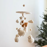 A charming corgi and sheep wool felt baby mobile hangs against a soft white wall, with a green pine tree partially visible on the right, creating a cozy nursery scene. The mobile features needle-felted figures suspended from a circular wooden frame, topped by a beige felt ball. A tan-and-white corgi is encircled by beige stars and clouds, while three fluffy wool felt sheep and more beige felt balls adorn the bottom.