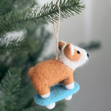 A wool felt corgi on a skateboard ornament is playfully displayed on a pine branch, set against a blurred white backdrop. This unique handcrafted felt ornament features a brown and white corgi with pointy ears, black eyes, and a short tail. The cheerful corgi stands confidently on a light blue skateboard with white wheels, complete with a jute string for hanging.