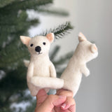 A handcrafted dire wolf felt finger puppet, perfect for festive storytelling, is held by a female hand against a blurred Christmas tree background. This unique wool felt puppet is primarily white, featuring small, pink-lined pointed ears, a black nose, and black eyes, highlighting its detailed design.