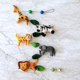 A vibrant wool felt African safari animal garland is playfully displayed on a white marble surface. This handmade felt garland features five detailed 3D animals: a brown-spotted giraffe, an orange-striped tiger, a black and white striped zebra, a gray elephant, and a lion with a brown mane. Green felt leaves and blue and green felt balls separate the animals along a thin black string, ending with a small copper bell.