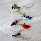 A vibrant felt American bird garland, featuring a green-winged hummingbird, a blue jay, a red cardinal, and a chickadee, is displayed diagonally on a light, marbled surface. The felt birds are connected by a wooden frame interspersed with felt leaves.